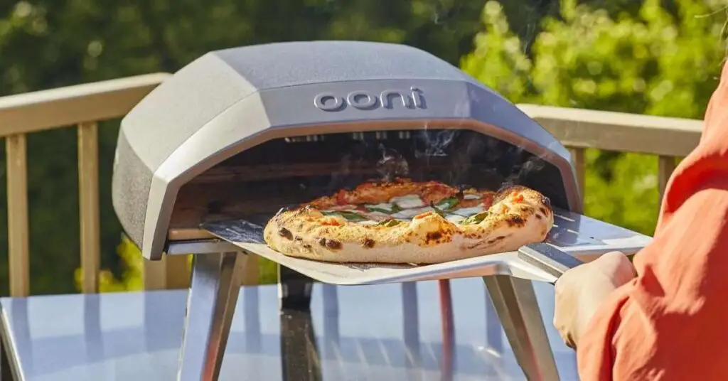 An Ooni Koda 12 propane powered pizza oven with a person pulling a pizza out from it using a pizza peel.