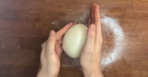 Two hands rolling a pizza dough ball on a brown table.