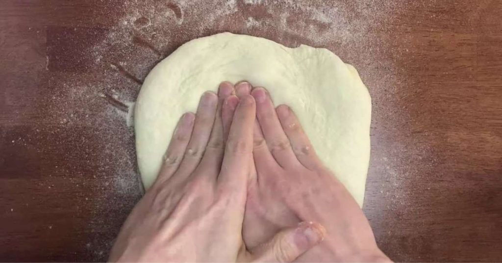 Two hands shaping homemade pizza dough on a table sprinkled with semolina flour to prevent sticking.