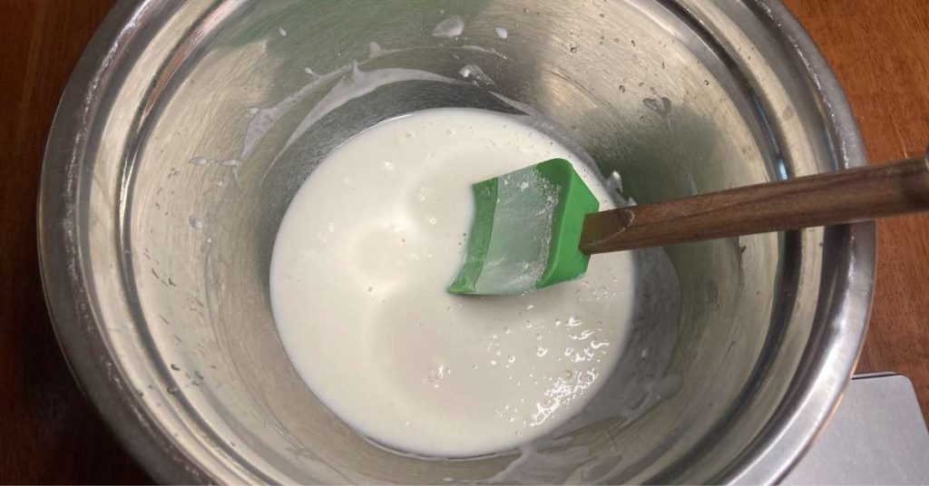 A green spatula sitting in a mixing bowl with water, yeast, and sugar.