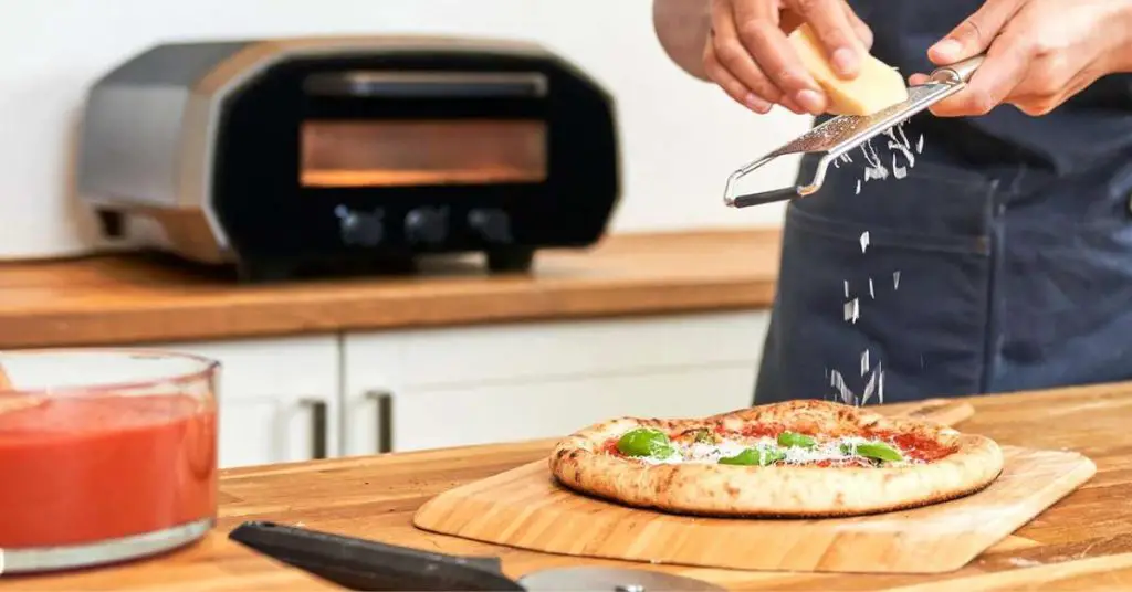 a chef grating cheese over a pizza made in the Ooni Volt 12 electric indoor pizza oven