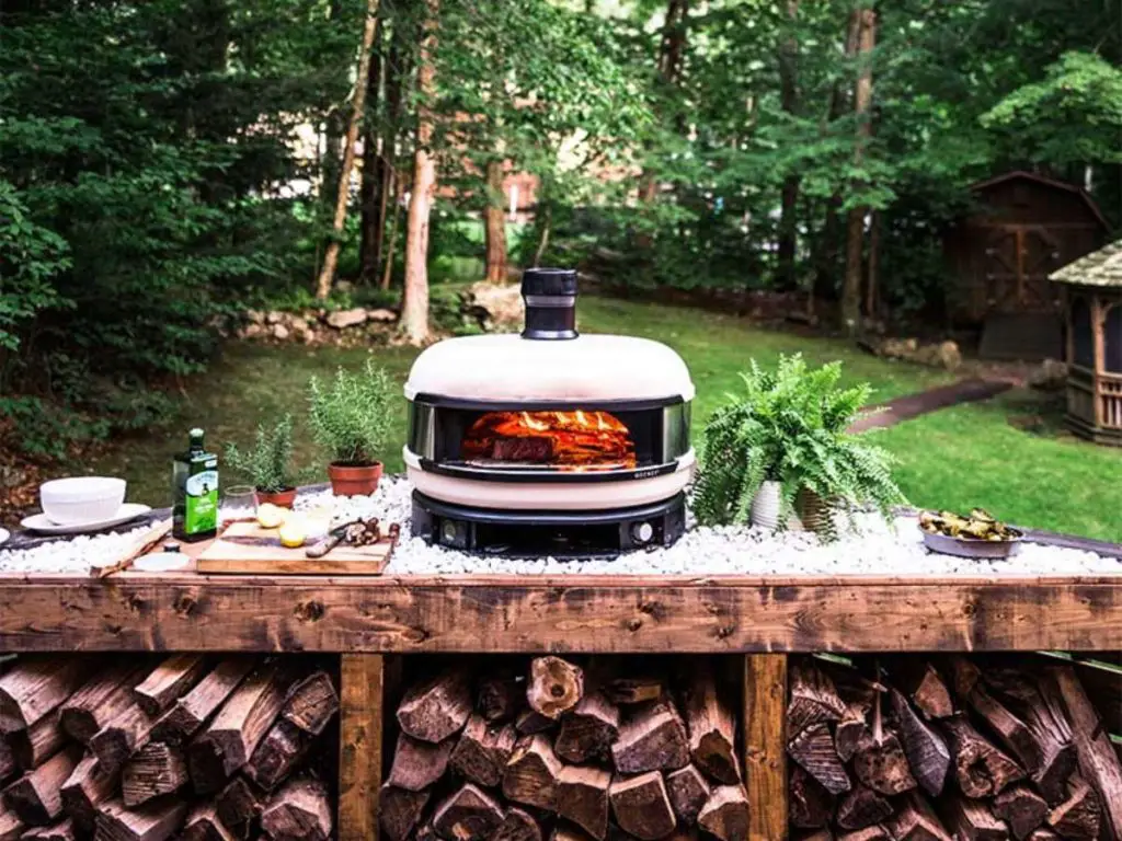 A white colored Gozney Dome pizza oven in a spacious backyard alongside firewood.