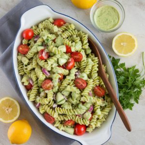 Creamy Avocado Pasta Salad Recipe That Will Steal the Show at Any Gathering 7 "Photo taken from a top-down angle, using all-natural lighting, on a light gray granite countertop. The finished avocado pasta salad is plated on a large, white serving dish, garnished with fresh parsley and surrounded by halved lemons and a small dish of extra dressing on the side. A wooden spoon is laid across the edge of the plate for serving."
