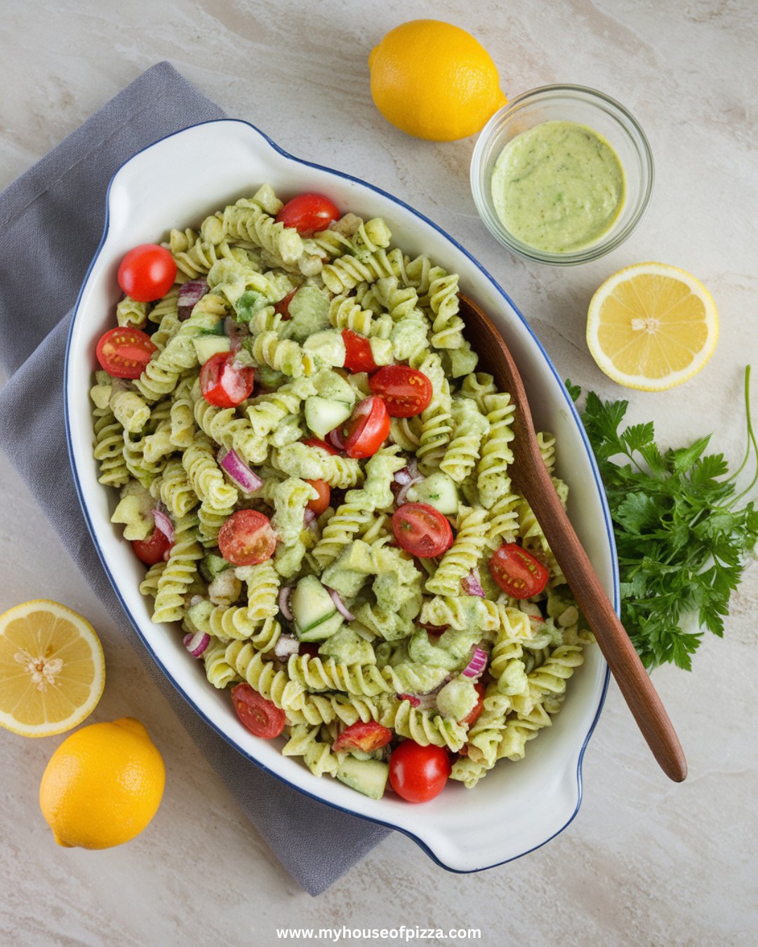 Creamy Avocado Pasta Salad Recipe That Will Steal the Show at Any Gathering 1 "Photo taken from a top-down angle, using all-natural lighting, on a light gray granite countertop. The finished avocado pasta salad is plated on a large, white serving dish, garnished with fresh parsley and surrounded by halved lemons and a small dish of extra dressing on the side. A wooden spoon is laid across the edge of the plate for serving."