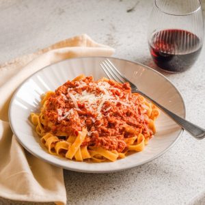 "Photo taken from a slightly side-angled view, using natural lighting, on a light gray granite countertop. A plate of freshly cooked tagliatelle is topped with a generous serving of Bolognese sauce. The dish is garnished with fresh basil leaves and a sprinkle of Parmesan cheese. A fork rests on the plate, with a glass of red wine and a cream-colored napkin casually placed nearby for a cozy, inviting setting."