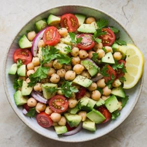 Fresh & Creamy Avocado Chickpea Salad Recipe 7 A photo taken from a top-down angle, using all-natural lighting. The completed avocado chickpea salad with chopped cucumber, halved cherry tomatoes, red onion, parsley, and drained chickpeas is plated in a shallow white bowl on a light gray granite countertop. The salad is garnished with a light sprinkle of parsley and black pepper, served with a lemon wedge on the side.