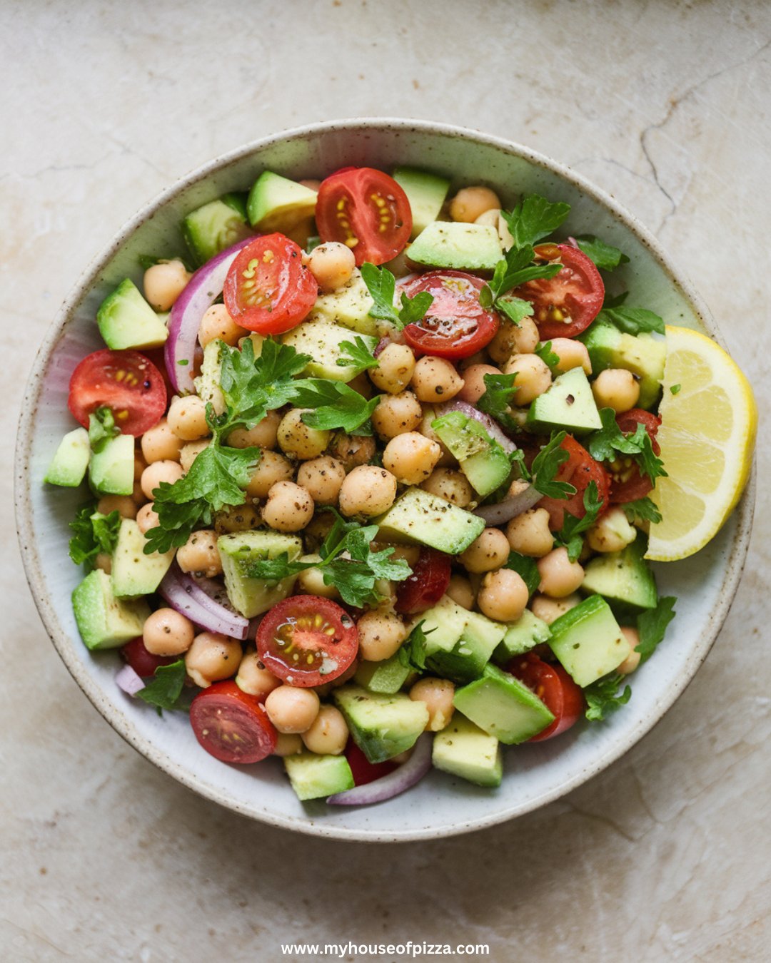 Fresh & Creamy Avocado Chickpea Salad Recipe 1 A photo taken from a top-down angle, using all-natural lighting. The completed avocado chickpea salad with chopped cucumber, halved cherry tomatoes, red onion, parsley, and drained chickpeas is plated in a shallow white bowl on a light gray granite countertop. The salad is garnished with a light sprinkle of parsley and black pepper, served with a lemon wedge on the side.