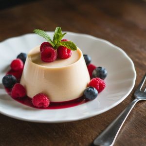 Creamy panna cotta topped with fresh raspberries and blueberries, garnished with mint leaves, elegantly presented on a white plate with a rustic wooden table background.
