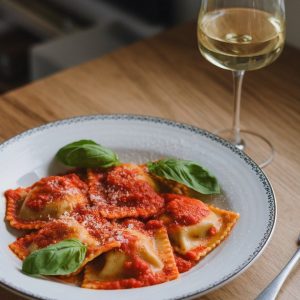 Creamy Spinach and Cheese Ravioli Recipe: Homemade Italian Comfort Food 7 A plate of spinach and cheese ravioli topped with rich tomato sauce, garnished with fresh basil leaves, served alongside a glass of white wine on a wooden table.