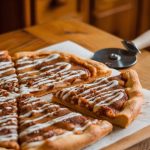 Homemade cinnamon sugar pizza topped with creamy icing drizzle on a wooden board, accompanied by a pizza cutter in a rustic kitchen setting.
