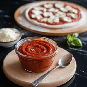 Copycat Pizza Hut Sauce Recipe: Make Your Favorite Takeout Pizza Sauce At Home 7 Homemade pizza sauce in a glass jar on a wooden board, with a spoon beside it, fresh basil leaves, and a partially prepared pizza topped with cheese and tomato sauce in the background on a black marble surface.