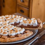 Gooey s'mores dessert pizza topped with toasted marshmallows, melted chocolate, and graham cracker pieces on a wooden table.