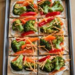 Colorful cold veggie pizza on a rectangular baking sheet topped with fresh broccoli, red bell peppers, shredded carrots, and creamy cheese spread on a crispy crust.