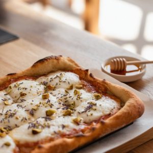 Honey Lavender Goat Cheese Flatbread Pizza – Elegant and Simple 7 Golden-brown flatbread pizza topped with melted cheese, sprinkled with crushed pistachios and lavender, sits on a wooden table beside a small dish of honey.
