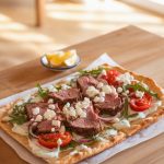 Savory lamb flatbread pizza topped with fresh arugula, juicy cherry tomatoes, creamy feta cheese, and a drizzle of white sauce, served on a wooden table with lemon wedges.