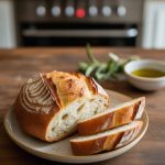 A beautifully baked rustic loaf of Italian bread, sliced to showcase its crusty exterior and soft, airy interior, perfect for dipping or serving alongside pasta dishes, presented on a neutral plate with a hint of greenery and olive oil in the background.
