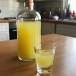 Refreshing homemade lemonade served in a clear shot glass alongside a glass bottle filled with yellow lemonade, set against a well-lit kitchen background.