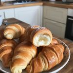 Freshly baked, golden-brown croissants arranged on a plate, showcasing their flaky texture and a partially visible creamy filling, set against a cozy kitchen backdrop. Perfect for a delightful breakfast or snack.