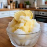 A refreshing bowl of homemade lemon sorbetto, featuring a fluffy, icy texture garnished with two lemon slices, set against a cozy kitchen backdrop, perfect for capturing the essence of Italian frozen desserts and summer treats.