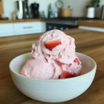 Delicious homemade strawberry ice cream served in a white bowl, featuring creamy pink scoops topped with fresh strawberry pieces, set against a modern kitchen backdrop. Perfect for summer desserts or special occasions.